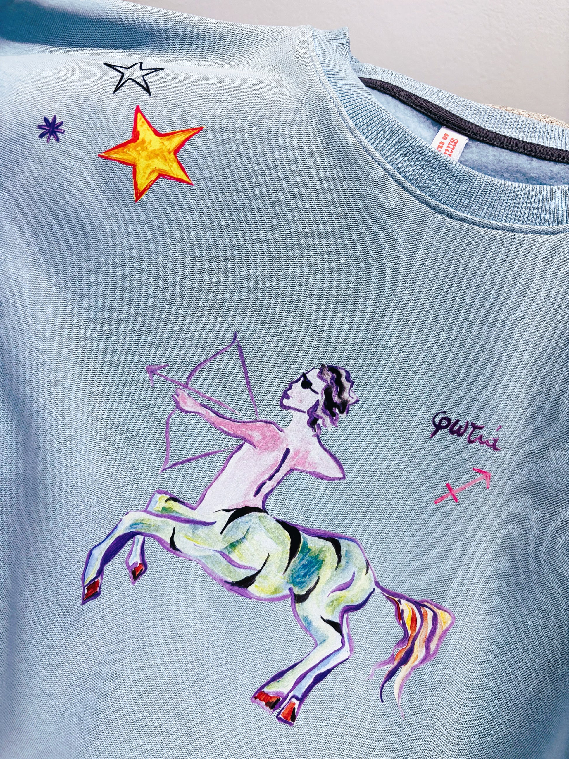 100% βαμβακερό φούτερ ελληνικής ραφής με πολύχρωμα τυπώματα μπροστά, πίσω και στα δύο μανίκια με θέμα το ζώδιο του Τοξότη σε γαλάζιο χρώμα.